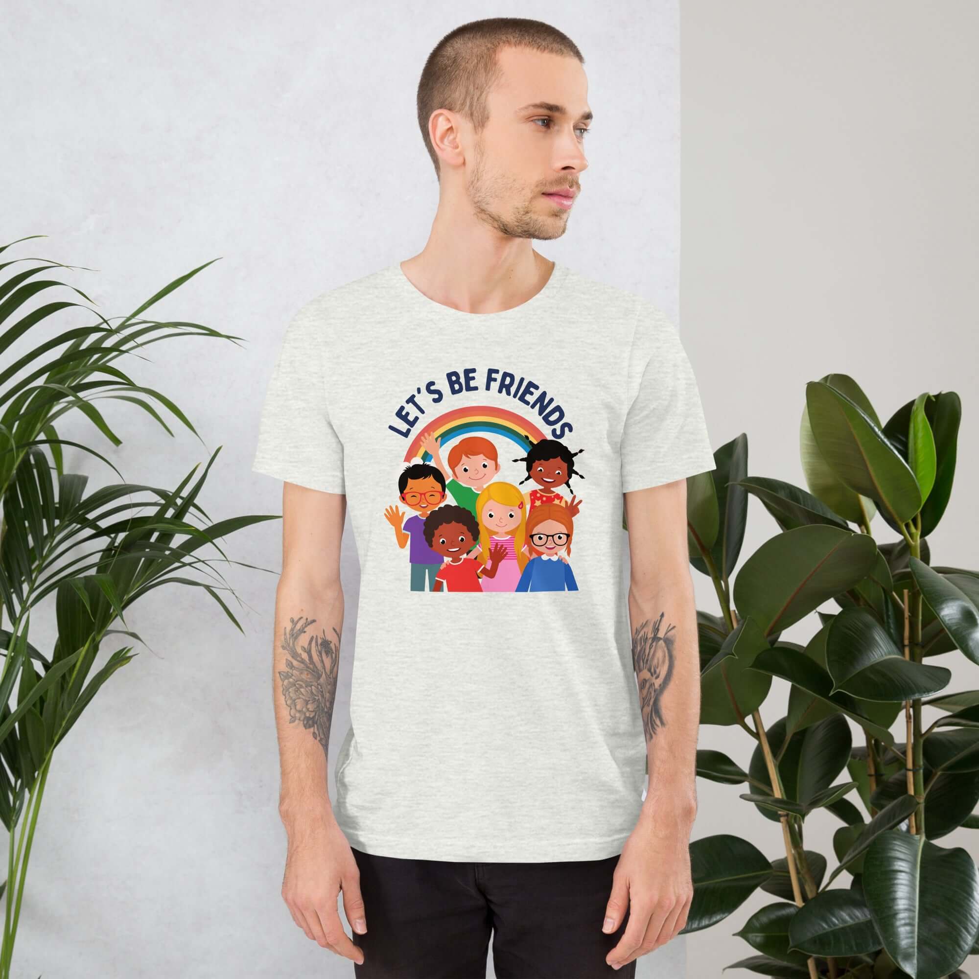 Man wearing a silver Milk Sandwich t-shirt featuring a colorful illustration of six diverse cartoon kids smiling under a rainbow with the text "LET'S BE FRIENDS" standing indoors next to large green plants.