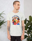 Man wearing a silver Milk Sandwich t-shirt featuring a colorful illustration of six diverse cartoon kids smiling under a rainbow with the text "LET'S BE FRIENDS" standing indoors next to large green plants.