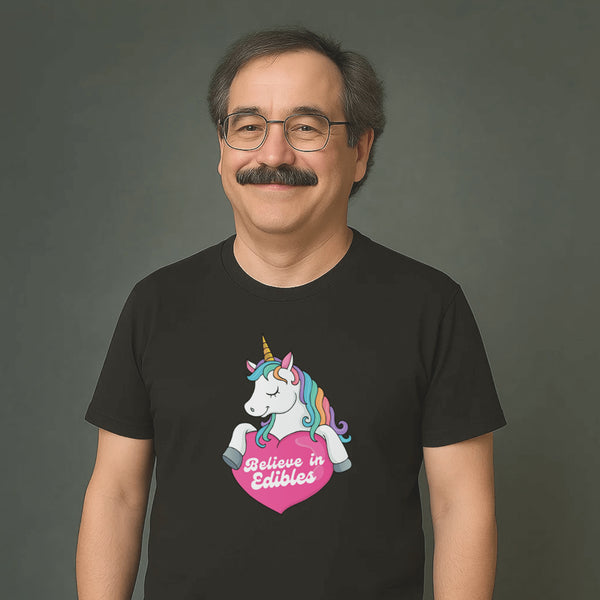 Smiling man with glasses and mustache wearing a black t-shirt with a colorful unicorn graphic and the text “Believe in Edibles” inside a pink heart.