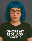 Woman with blue hair and glasses wearing a black “IGNORE MY RED FLAGS I’M WORTH IT” t-shirt, looking cheeky.