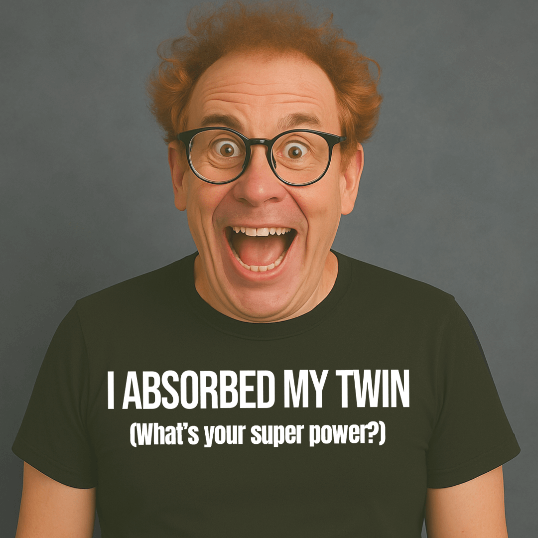 Excited man with glasses wearing black “I ABSORBED MY TWIN (What’s your superpower?)” t-shirt, smiling wide.