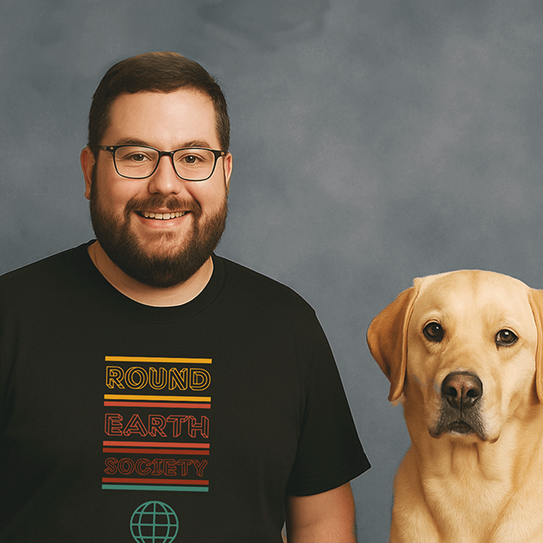 Smiling man with glasses and a beard wearing a black t-shirt that says ‘Round Earth Society’ in colorful text, standing next to a yellow Labrador Retriever dog