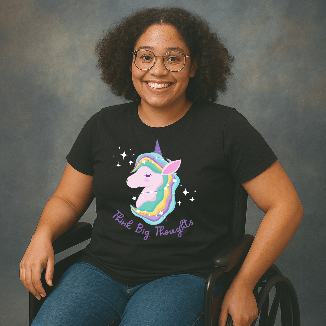 Smiling woman with curly hair and glasses sitting in a wheelchair, wearing a black t-shirt with a colorful unicorn graphic and the text ‘Think Big Thoughts