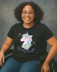 Smiling woman with curly hair and glasses sitting in a wheelchair, wearing a black t-shirt with a colorful unicorn graphic and the text ‘Think Big Thoughts