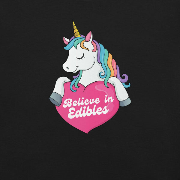 Close-up of a colorful unicorn graphic holding a pink heart with the text “Believe in Edibles” on a black fabric background.