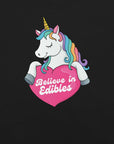 Close-up of a colorful unicorn graphic holding a pink heart with the text “Believe in Edibles” on a black fabric background.