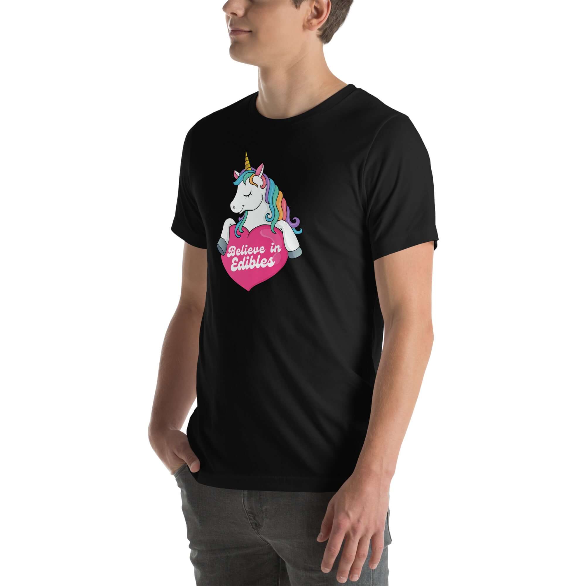 Young man wearing a black t-shirt with a colorful unicorn holding a pink heart that says “Believe in Edibles,” standing with hands in pockets, side view.