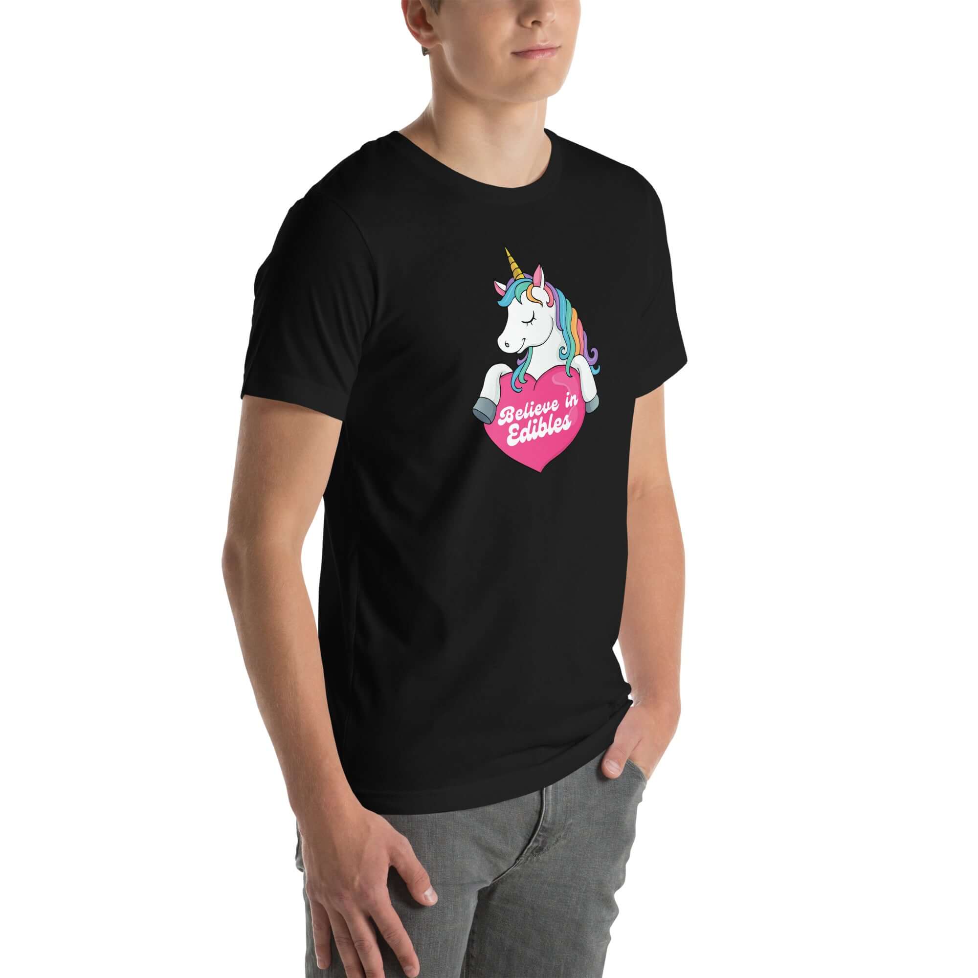 Man wearing a black t-shirt with a colorful unicorn graphic holding a pink heart that says “Believe in Edibles,” standing with hands in pockets, front and side view.