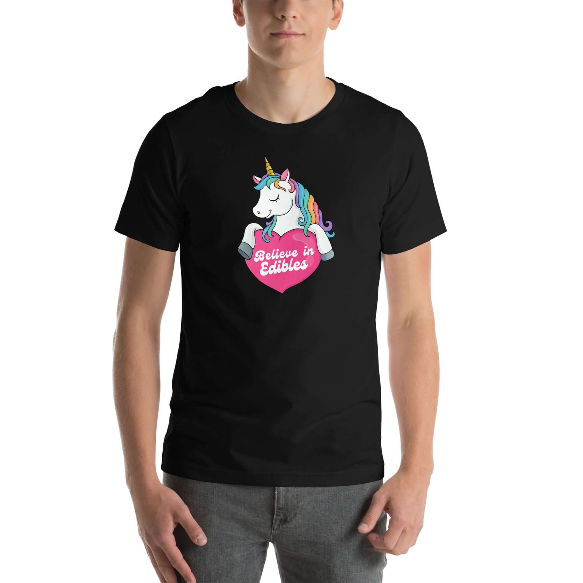 Man wearing a black t-shirt with a colorful unicorn holding a pink heart that says “Believe in Edibles,” standing facing forward with one hand relaxed and the other in his pocket.          