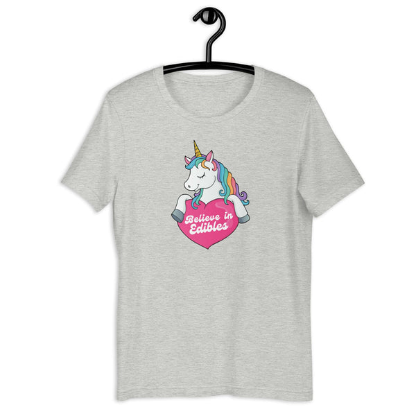Light grey t-shirt on a hanger with a colorful unicorn graphic above a pink heart that says “Believe in Edibles.”