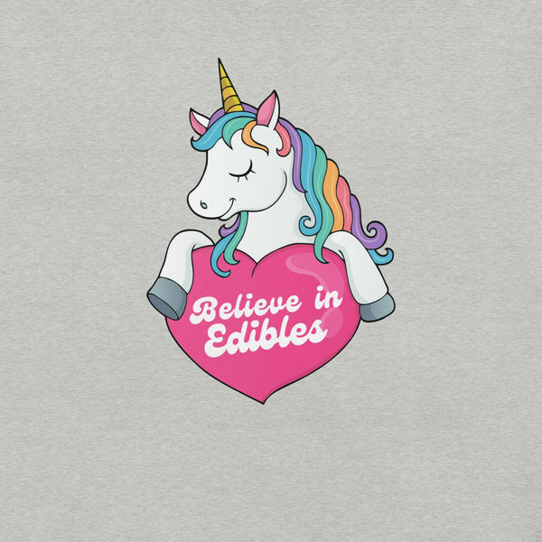 Close-up of a colorful unicorn graphic holding a pink heart with the text “Believe in Edibles” on a light grey fabric background.