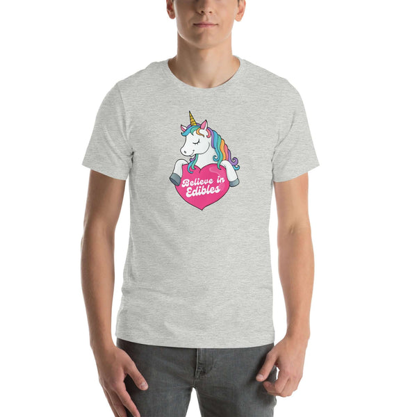 Man wearing a light grey t-shirt with a colorful unicorn holding a pink heart that says “Believe in Edibles,” standing facing forward with one hand relaxed and the other in his pocket.          