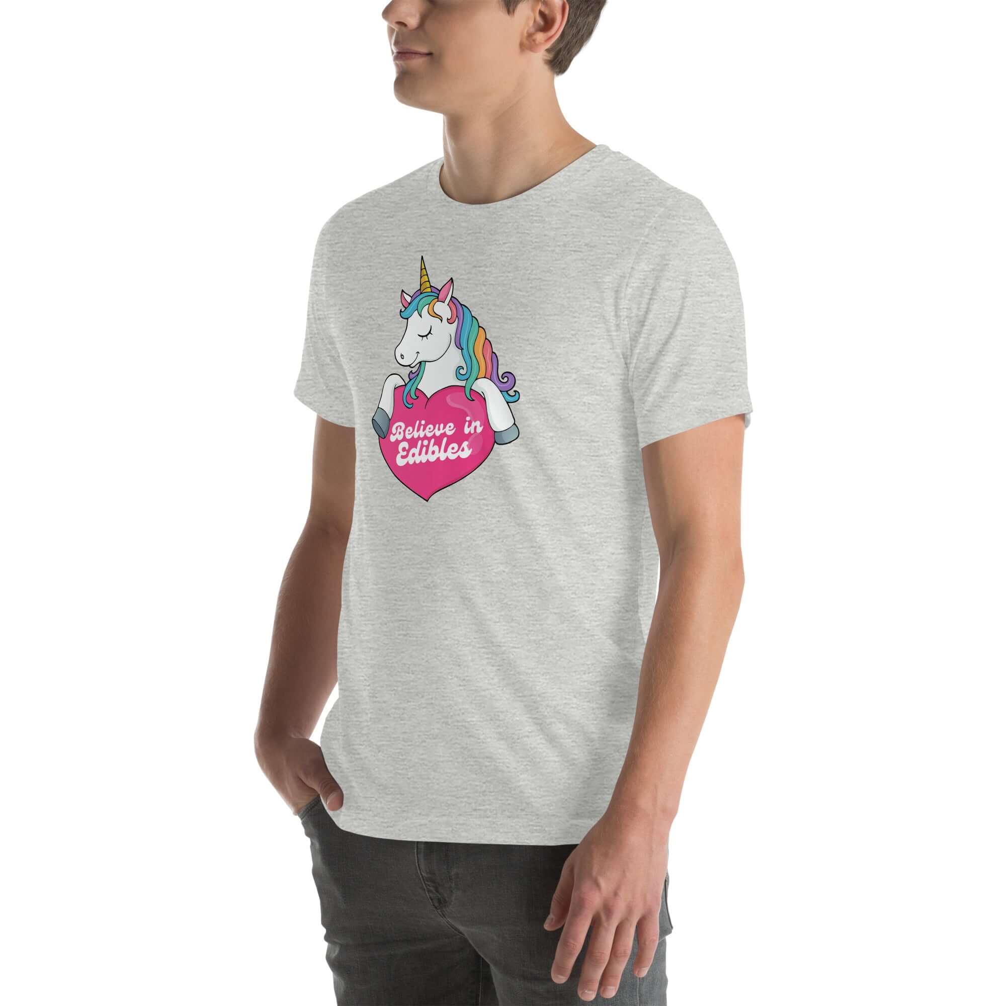 Young man wearing a light grey t-shirt with a colorful unicorn holding a pink heart that says “Believe in Edibles,” standing with hands in pockets, side view.