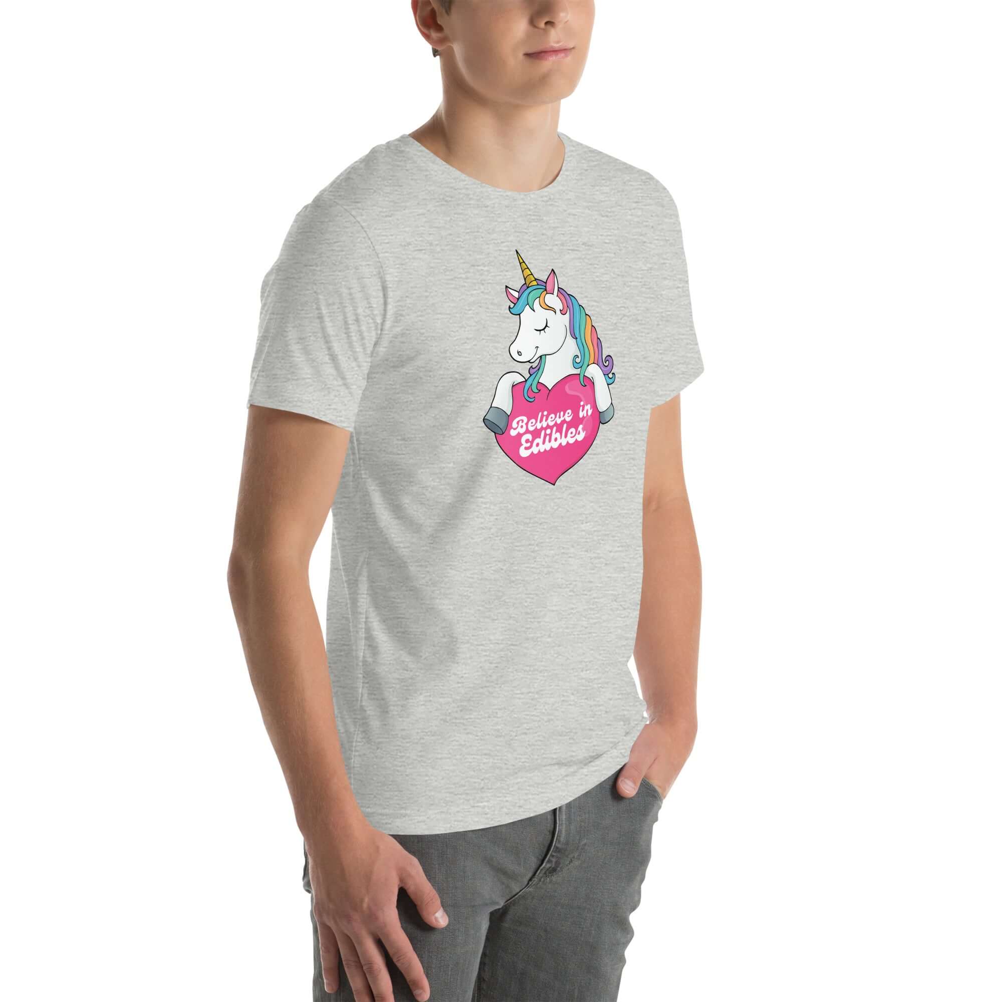 Young man wearing a light grey t-shirt with a colorful unicorn holding a pink heart that says “Believe in Edibles,” standing with hands in pockets, side view.