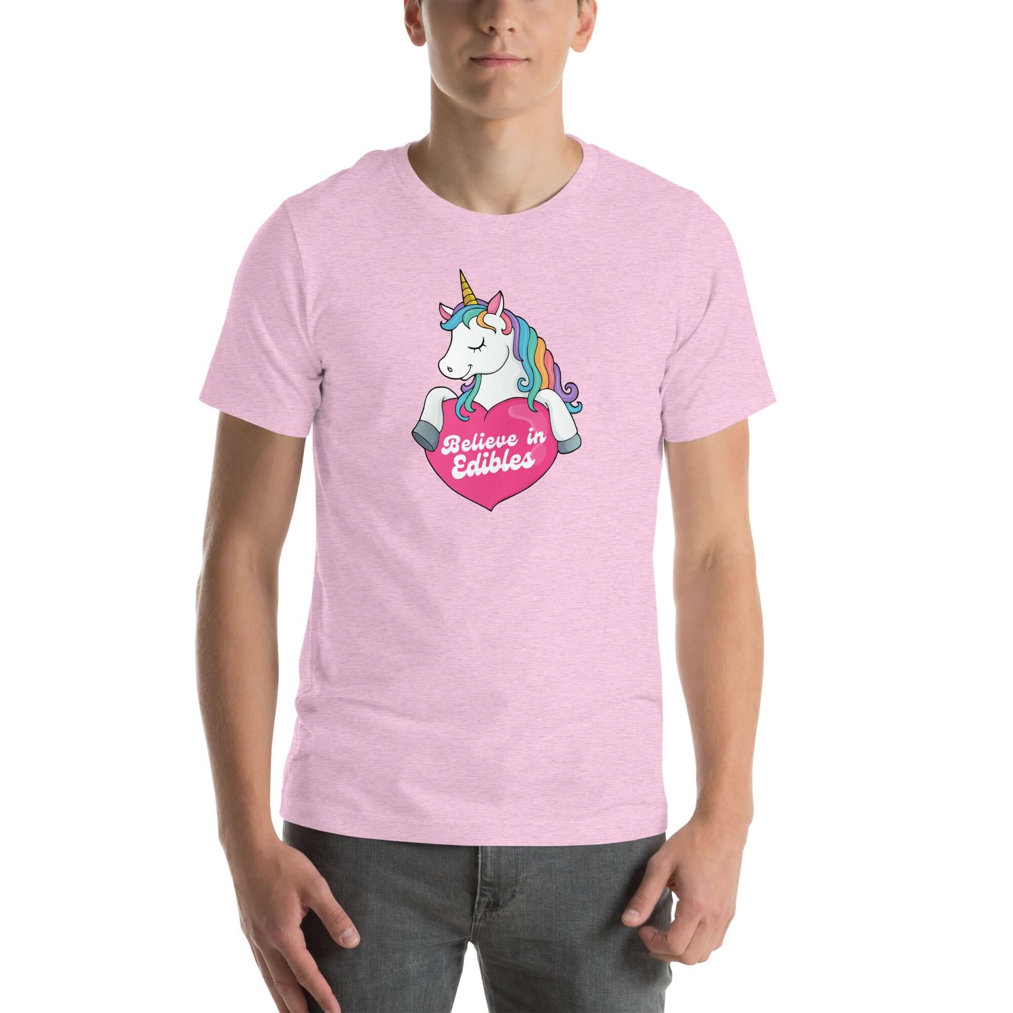 Man wearing a lilac t-shirt with a colorful unicorn holding a pink heart that says “Believe in Edibles,” standing facing forward with one hand relaxed and the other in his pocket.          