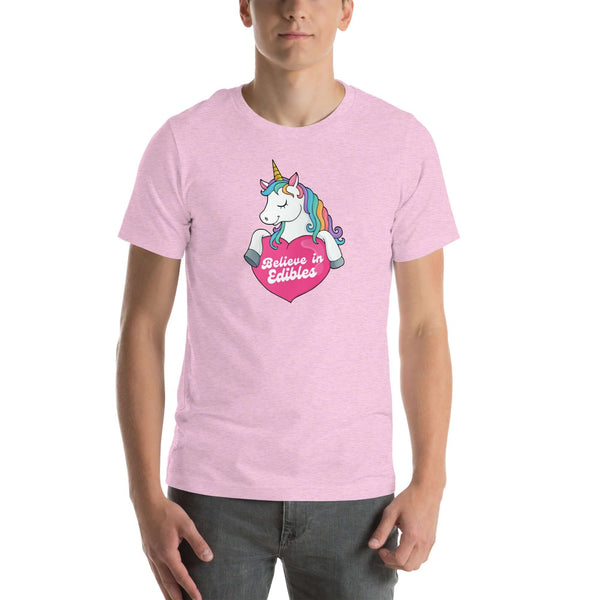 Man wearing a lilac t-shirt with a colorful unicorn holding a pink heart that says “Believe in Edibles,” standing facing forward with one hand relaxed and the other in his pocket.          