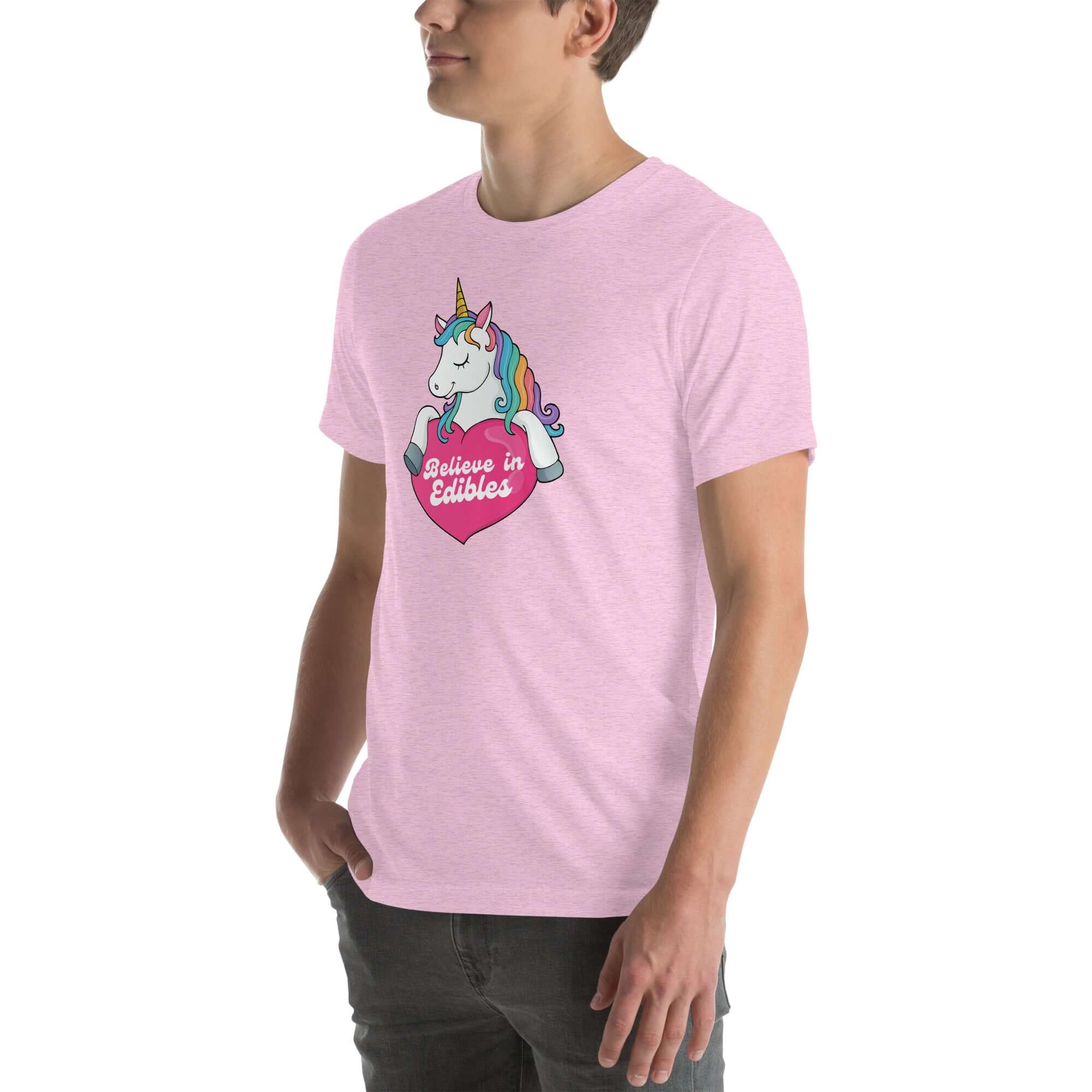 Young man wearing a lilac t-shirt with a colorful unicorn holding a pink heart that says “Believe in Edibles,” standing with hands in pockets, side view.