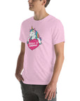 Young man wearing a lilac t-shirt with a colorful unicorn holding a pink heart that says “Believe in Edibles,” standing with hands in pockets, side view.