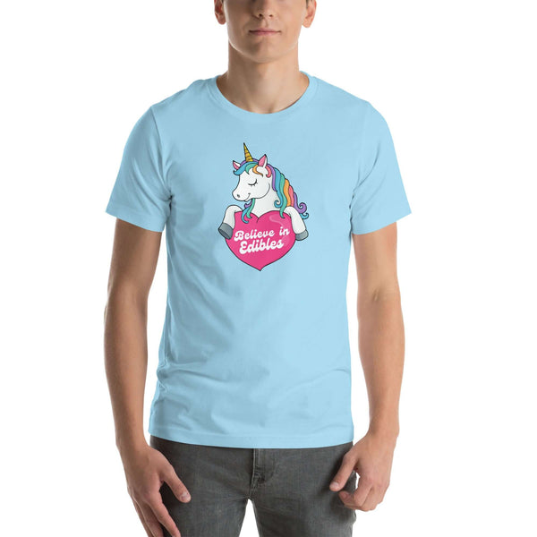 Man wearing a ocean blue t-shirt with a colorful unicorn holding a pink heart that says “Believe in Edibles,” standing facing forward with one hand relaxed and the other in his pocket.          