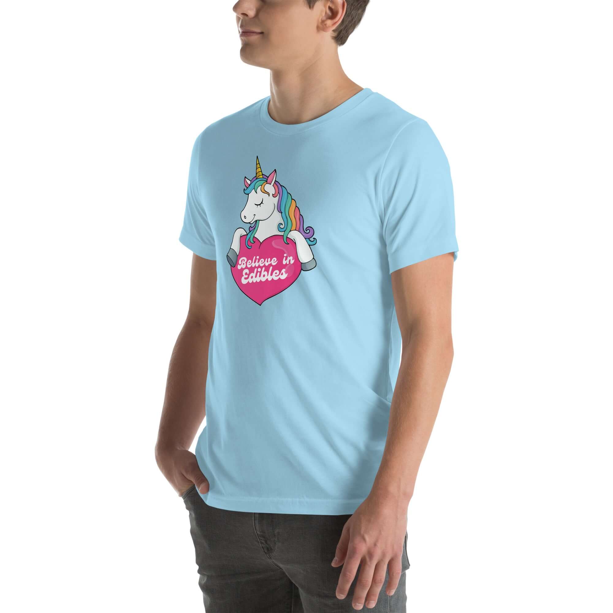 Young man wearing a ocean blue t-shirt with a colorful unicorn holding a pink heart that says “Believe in Edibles,” standing with hands in pockets, side view.
