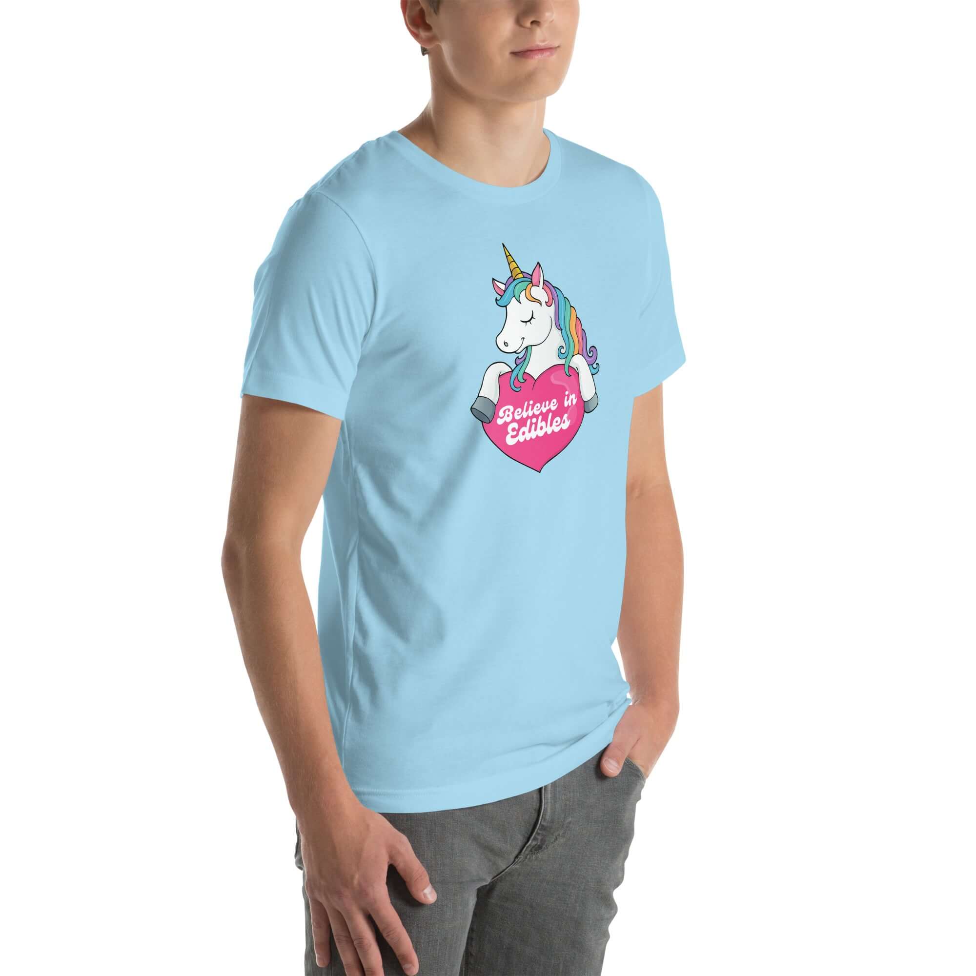 Young man wearing a ocean blue t-shirt with a colorful unicorn holding a pink heart that says “Believe in Edibles,” standing with hands in pockets, side view.