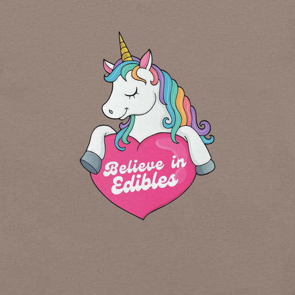 Close-up of a colorful unicorn graphic holding a pink heart with the text “Believe in Edibles” on a pebble fabric background.