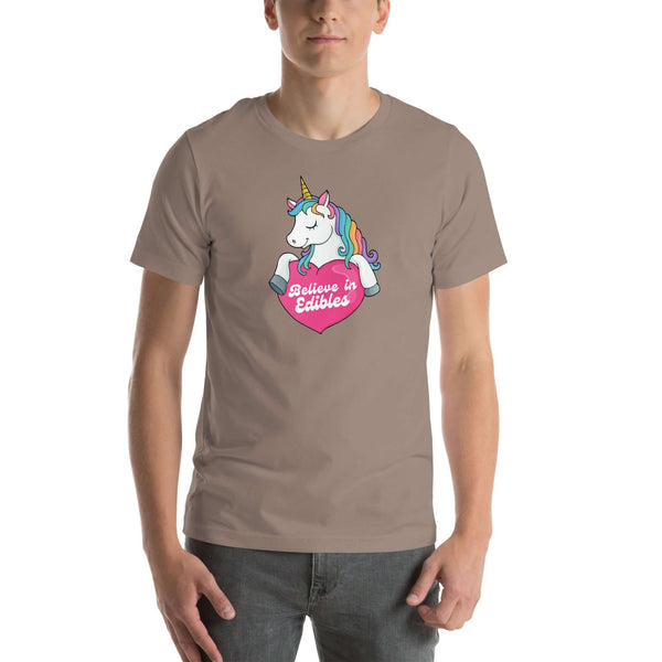 Man wearing a pebble t-shirt with a colorful unicorn holding a pink heart that says “Believe in Edibles,” standing facing forward with one hand relaxed and the other in his pocket.          