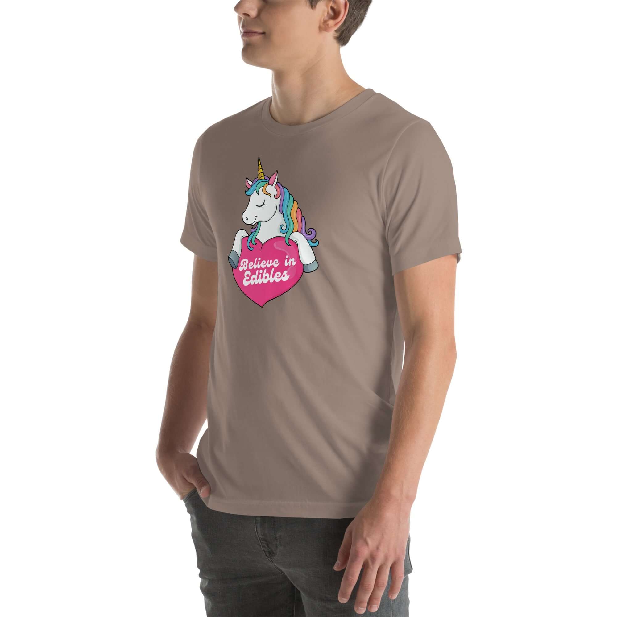 Young man wearing a pebble t-shirt with a colorful unicorn holding a pink heart that says “Believe in Edibles,” standing with hands in pockets, side view.