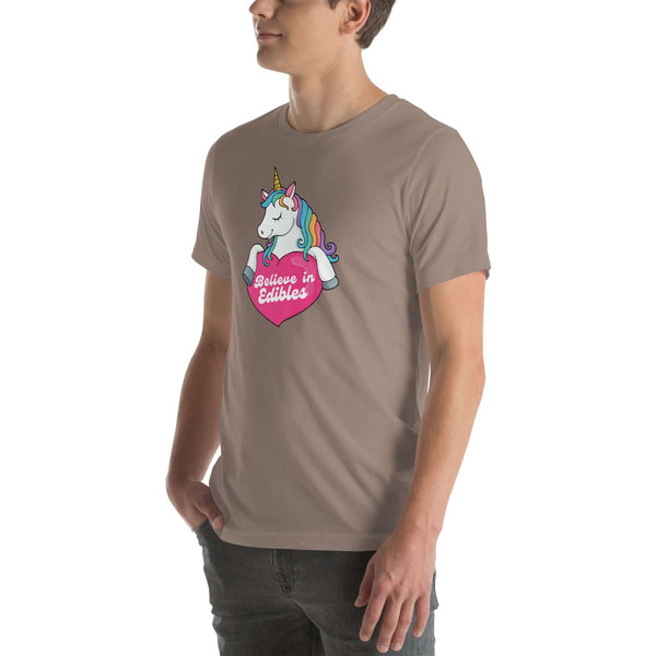 Young man wearing a pebble t-shirt with a colorful unicorn holding a pink heart that says “Believe in Edibles,” standing with hands in pockets, side view.