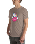 Young man wearing a pebble t-shirt with a colorful unicorn holding a pink heart that says “Believe in Edibles,” standing with hands in pockets, side view.