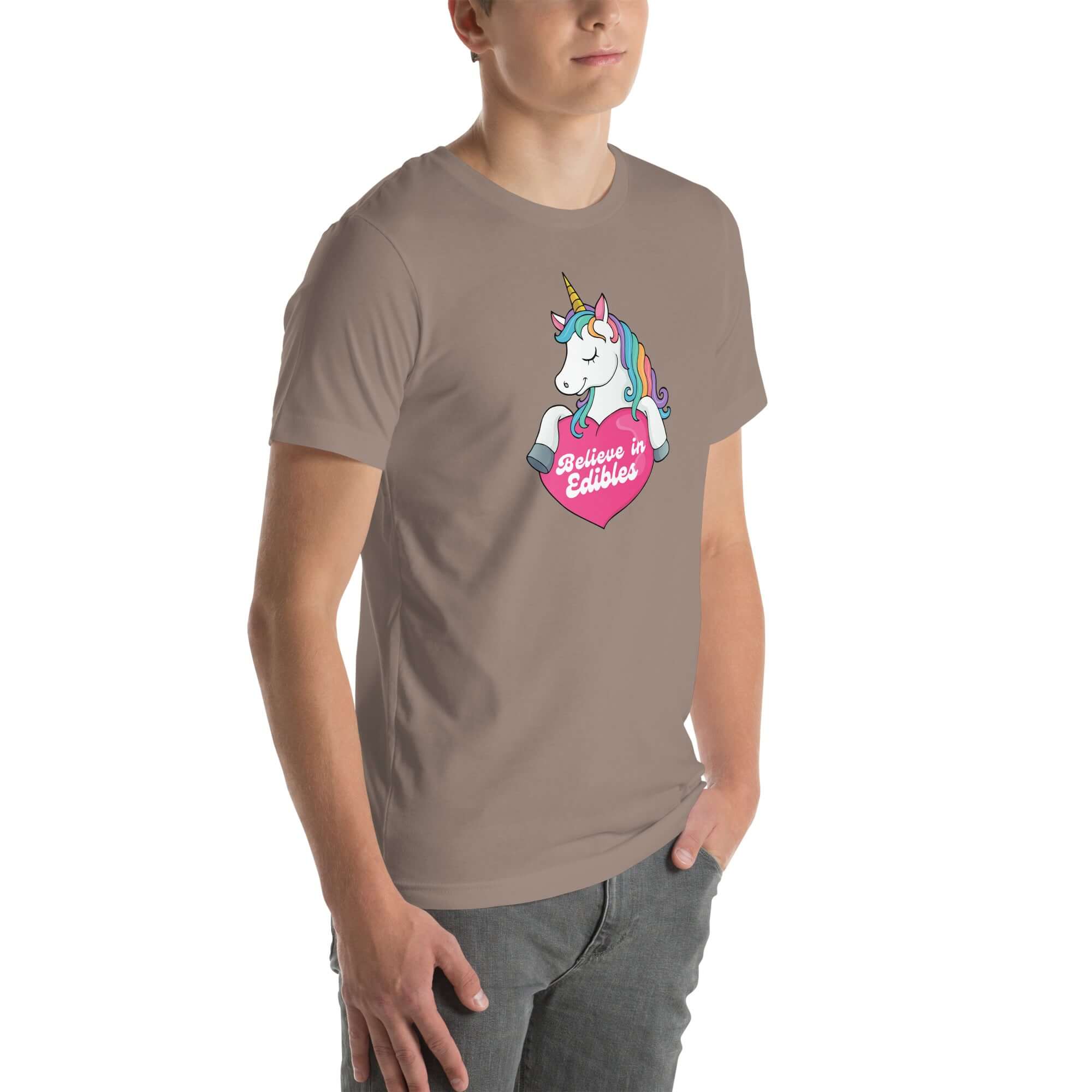 Young man wearing a pebble t-shirt with a colorful unicorn holding a pink heart that says “Believe in Edibles,” standing with hands in pockets, side view.