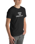 Young man wearing a black t-shirt with white text ‘HIGH ON CREATIVITY’, standing angled to the side with hands in pockets.