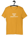 Mustard t-shirt on a hanger with the phrase “HIGH ON CREATIVITY” printed in white, centered on the chest.