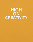 Close-up of mustard t-shirt fabric with the phrase “HIGH ON CREATIVITY” printed in bold white uppercase letters in three lines.