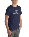Young man wearing a navy t-shirt with white text ‘HIGH ON CREATIVITY’, standing angled to the side with hands in pockets.