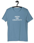 Steel Blue t-shirt on a hanger with the phrase “HIGH ON CREATIVITY” printed in white, centered on the chest.