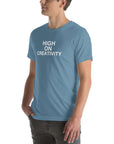 Man wearing a steel blue t-shirt with white text ‘HIGH ON CREATIVITY’, standing sideways with one hand in pocket