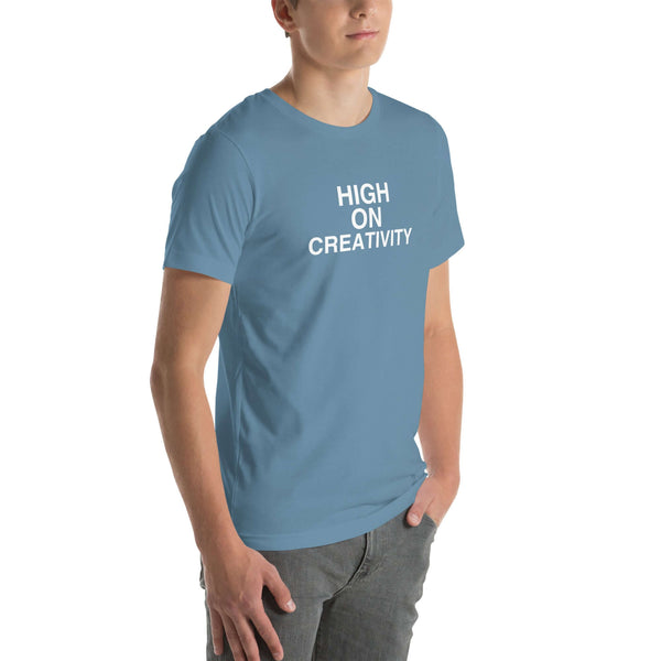 Young man wearing a steel blue t-shirt with white text ‘HIGH ON CREATIVITY’, standing angled to the side with hands in pockets.