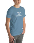 Young man wearing a steel blue t-shirt with white text ‘HIGH ON CREATIVITY’, standing angled to the side with hands in pockets.