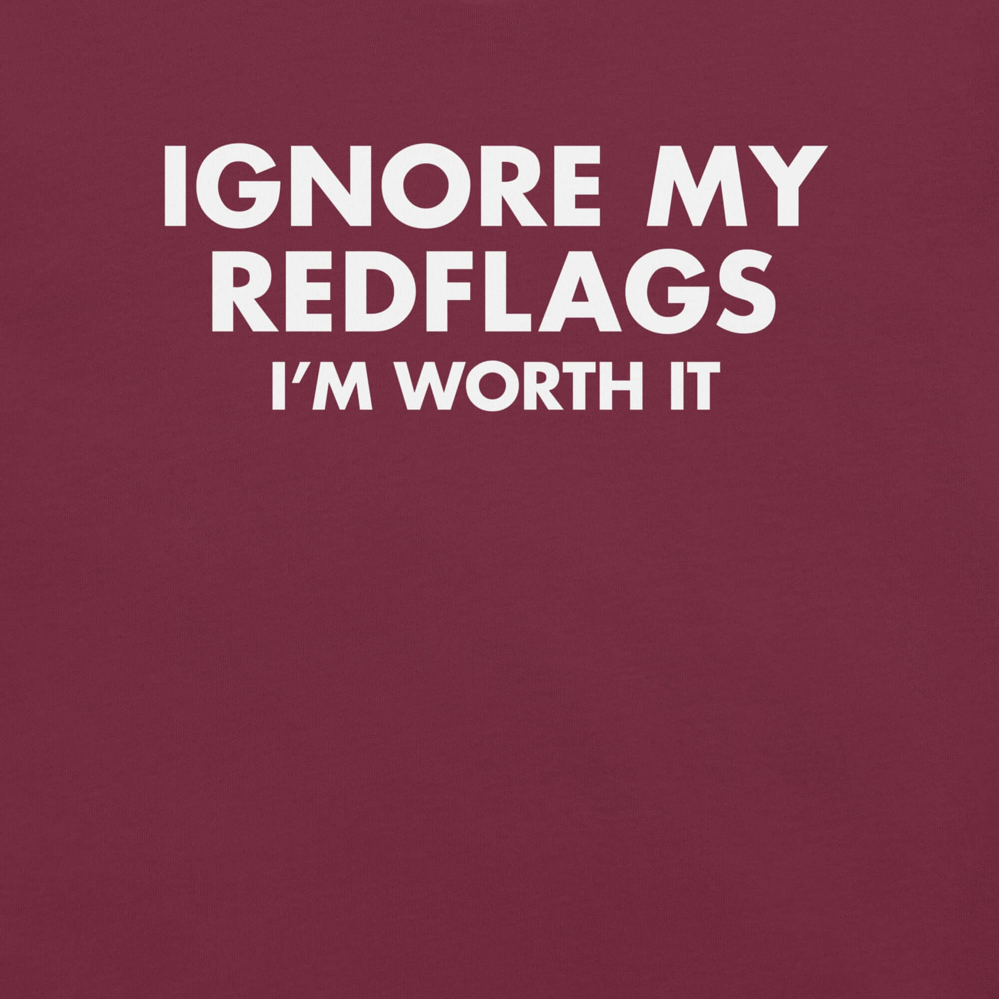 Close-up of maroon t-shirt with bold white text “IGNORE MY RED FLAGS I’M WORTH IT” printed on the front.        