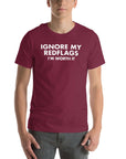 Man wearing maroon “IGNORE MY RED FLAGS I’M WORTH IT” t-shirt, standing casually with hands by pockets.