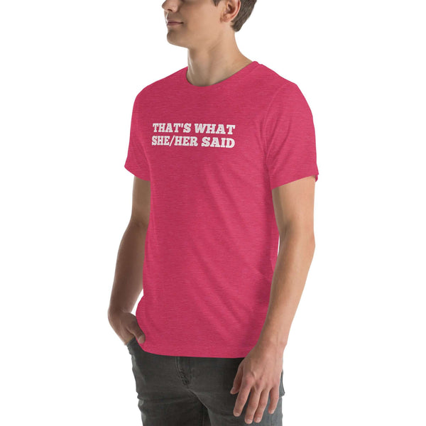 Man wearing a pink “THAT’S WHAT SHE/HER SAID” t-shirt, standing with hands in pockets, looking to the side.