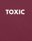 Man wearing maroon “TOXIC” t-shirt, standing with hands in pockets, looking slightly to the side.