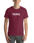 Man wearing maroon “TOXIC” t-shirt, standing straight with hands relaxed by pockets, facing forward.