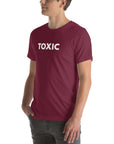 Man wearing maroon “TOXIC” t-shirt, standing sideways with hands in pockets, looking off to the side.