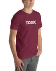 Man wearing maroon “TOXIC” t-shirt, standing with hands in pockets, looking slightly to the side.