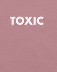 Man wearing orchid “TOXIC” t-shirt, standing with hands in pockets, looking slightly to the side.