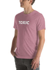 Man wearing orchid “TOXIC” t-shirt, standing sideways with hands in pockets, looking off to the side.