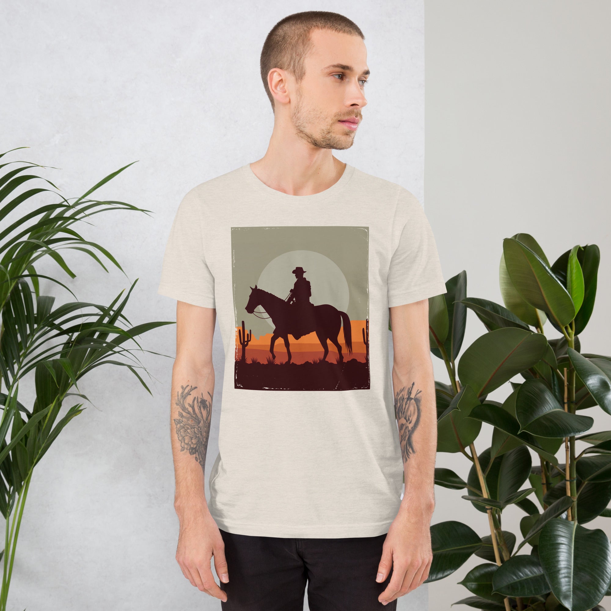 Unisex t-shirt featuring cowboy graphic with sunset, worn by model in stylish room with plants. Perfect blend of comfort and style.